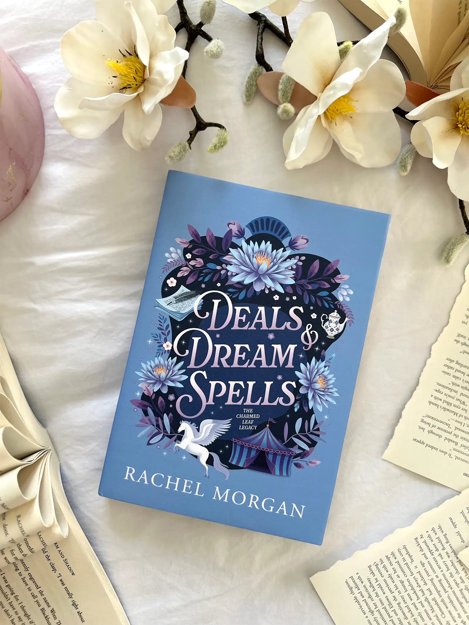 A flat lay of the Deals & Dream Spells special edition hardback, styled with flowers and loose book pages.